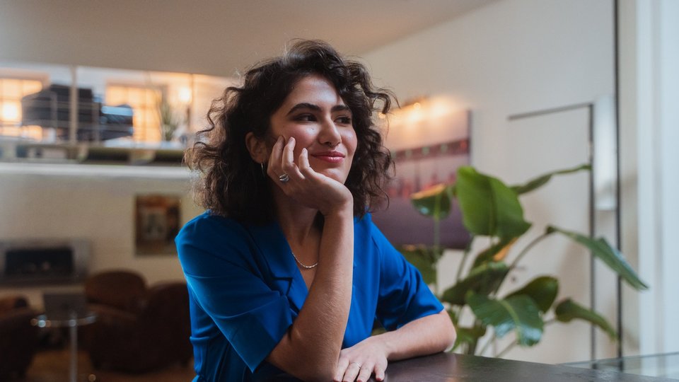 Young woman in a blue blouse sits at a table, gazing into the distance with a thoughtful smile.