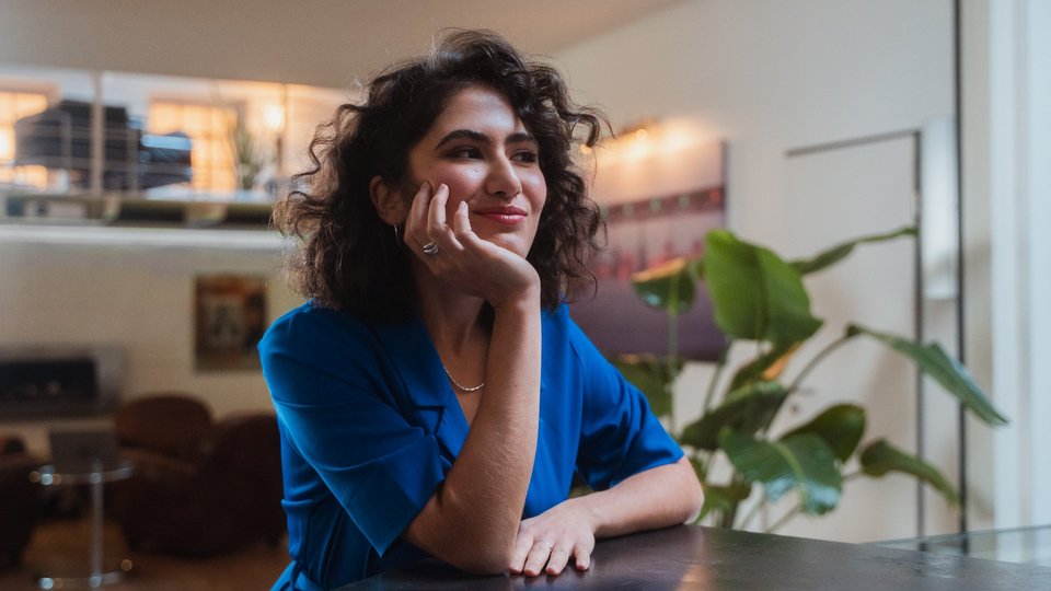 Junge Frau in blauer Bluse sitzt an einem Tisch und blickt nachdenklich grinsend in die Ferne.