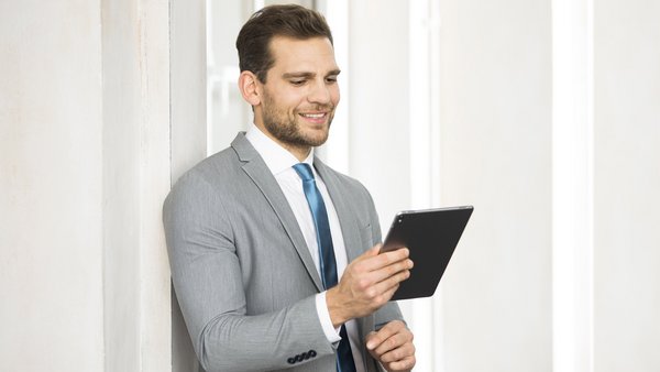 Geschäftsmann im Anzug hält ein Tablet und liest konzentriert, im Hintergrund lichtdurchflutetes Büro.