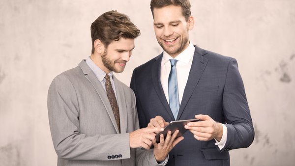 Zwei Männer in Anzügen schauen gemeinsam auf ein Tablet und lächeln.