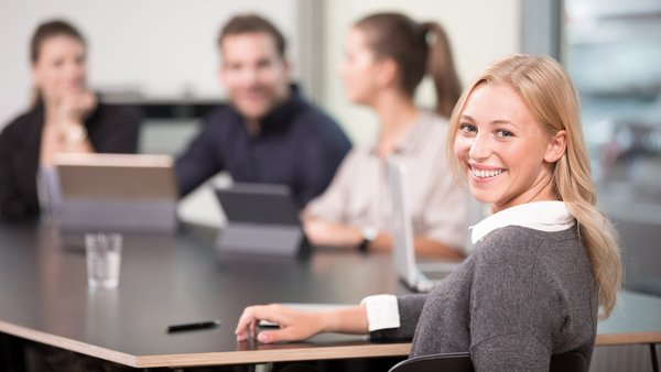 Frau am Konferenztisch lächelt in die Kamera, im Hintergrund weitere Personen bei einem Meeting.