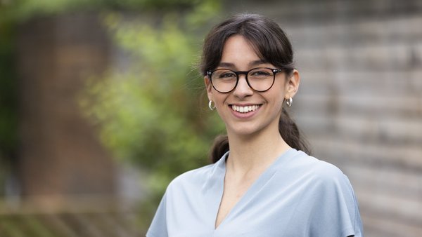 Junge Frau mit Brille lächelt in die Kamera, steht im Freien vor einem grünen Hintergrund.