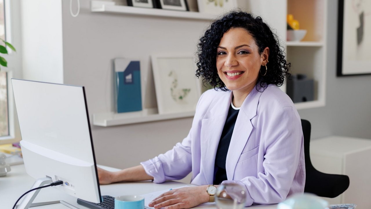 junge Frau lächelt und schaut über ihre Schulter. Dahinter sind zwei weitere Personen an einem Tisch zu sehen.