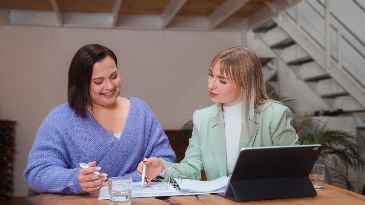 Two women sit at a table in front of a laptop and look at documents together.